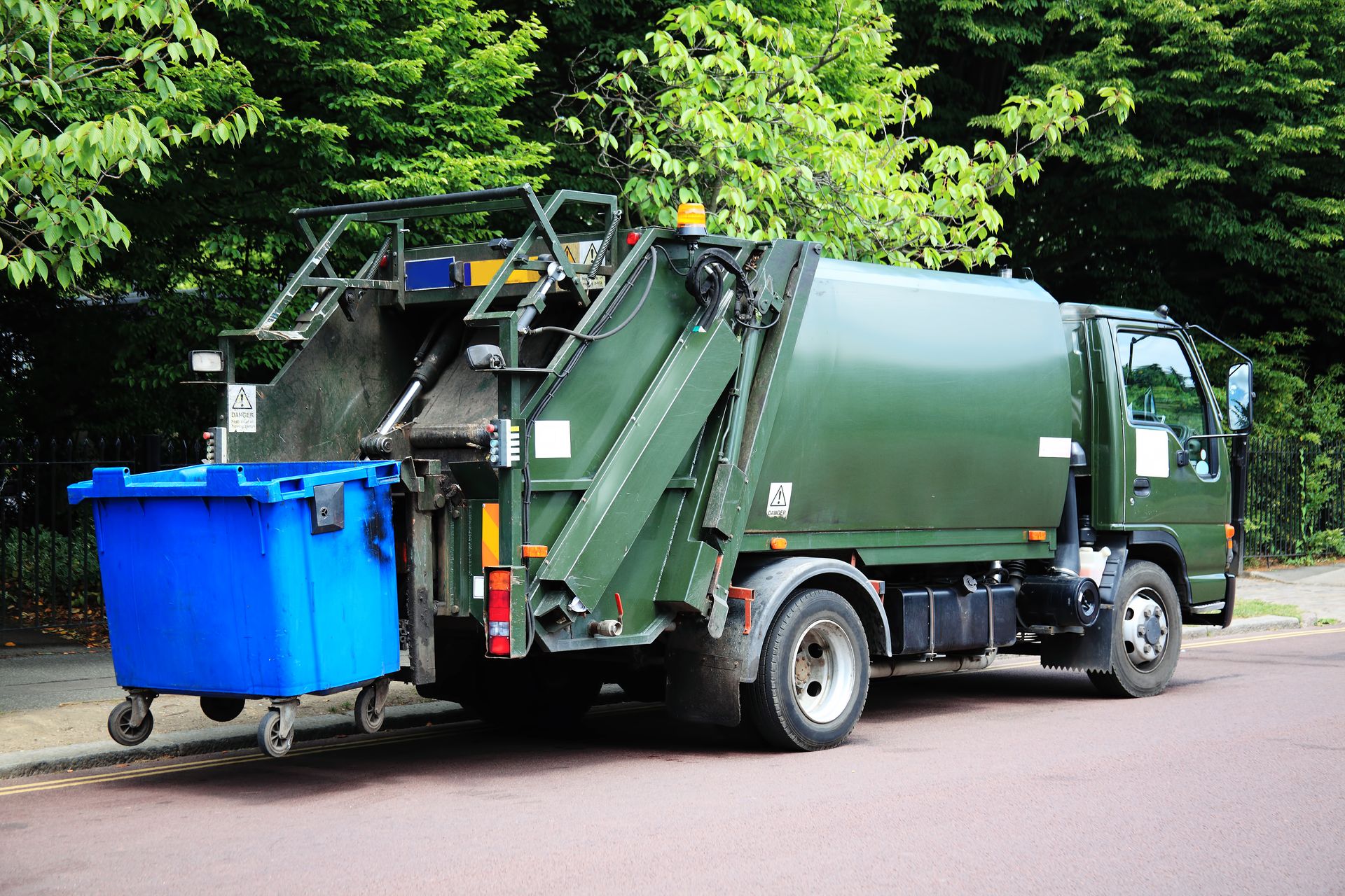 Green garbage truck with an elevated blue wheelie bin at the rear